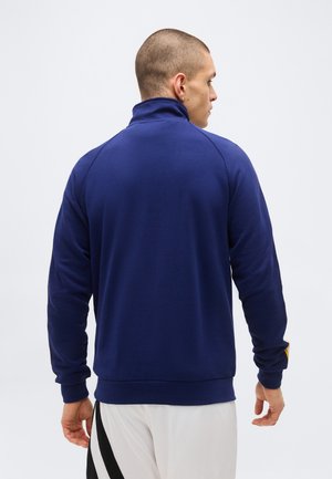 Mann mit kurzen Haaren, von hinten gesehen, trägt eine marineblaue Jacke mit langen Ärmeln und weiße Hosen mit schwarzen Streifen.