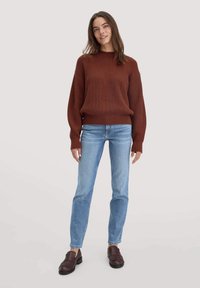 Rotbrauner, gerippter Strickpullover mit rundem Ausschnitt, kombiniert mit hellblauen, geraden Jeans und dunklen Slippern. Einfache, strukturierte Gestaltung.
