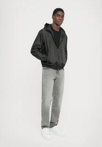 Svart lättviktsjacka med huva, strukturerat mönster, dragkedjestängning, kombinerad med ljusgrå jeans och vita sneakers.