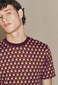 Tričko s krátkym rukávom a okrúhlym výstrihom s geometrickým vzorom v odtieňoch bordovej, krémovej a hnedej. Mäkká textúra a detail s rebrovaným golierom.