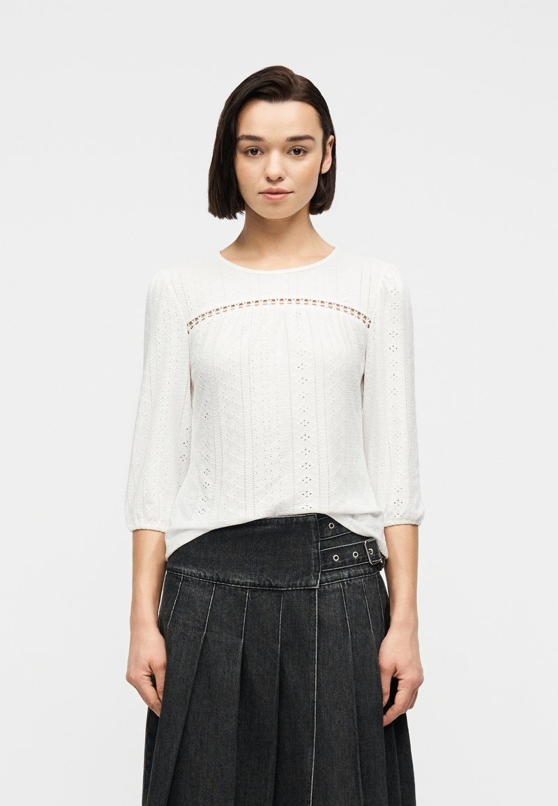 Γυναίκα με κοντά σκούρα μαλλιά, φορώντας λευκό μπλουζάκι με τρύπες και μανίκια 3/4, καθώς και μαύρη πλισέ φούστα ψηλής μέσης με λεπτομέρεια αγκράφας.