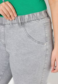 Pantalones de mezclilla gris con una cinturilla elástica acanalada, bolsillos laterales y una textura sutil. Mano descansando en la cintura para escala.