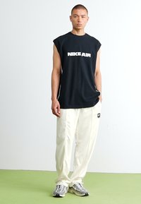Sort ærmeløs t-shirt med hvid "NIKE AIR" logo, parret med hvide træningsbukser og grå sneakers, stående mod en lys baggrund.