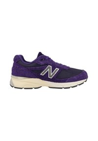 Zapatilla deportiva de color púrpura con una parte superior de textura suave y difusa, paneles de malla y una suela de color crema. Presenta un logotipo plateado en el lateral.