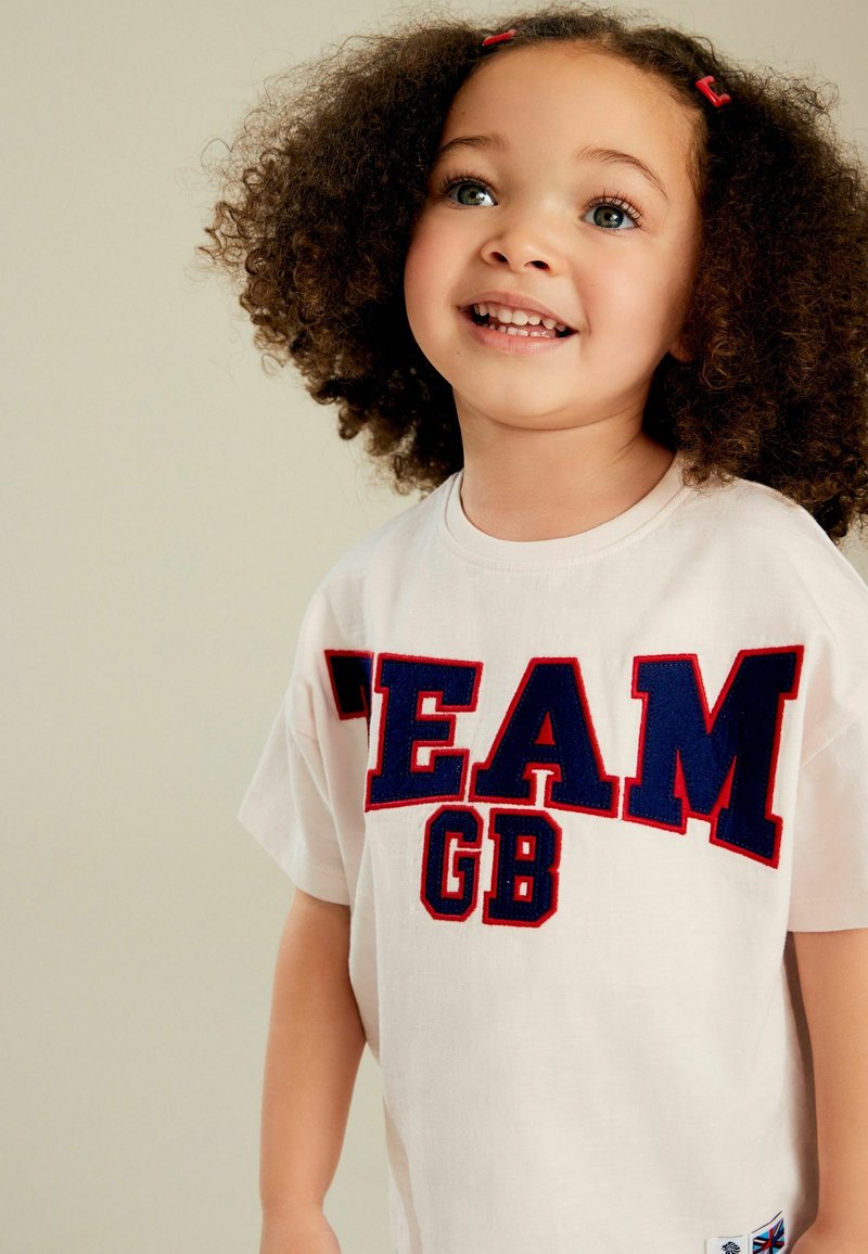 Cremfarbendes T-Shirt mit auffälligem navyblauen und roten "TEAM GB"-Schriftzug. Kurze Ärmel, lockere Passform und kleines Etikett mit Union Jack-Detail am Saum.