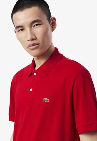 Röd polo-tröja i bomull, med en tvåknapps-krage och en grön broderad krokodil-logga på vänster bröst. Slät textur.