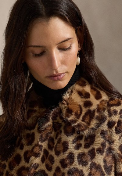 Kabát z umelej kožušiny s leopardím vzorom, zaobleným golierom a gombíkovými detailmi, nosený cez čierny rolák. Má mäkký, textúrovaný povrch.