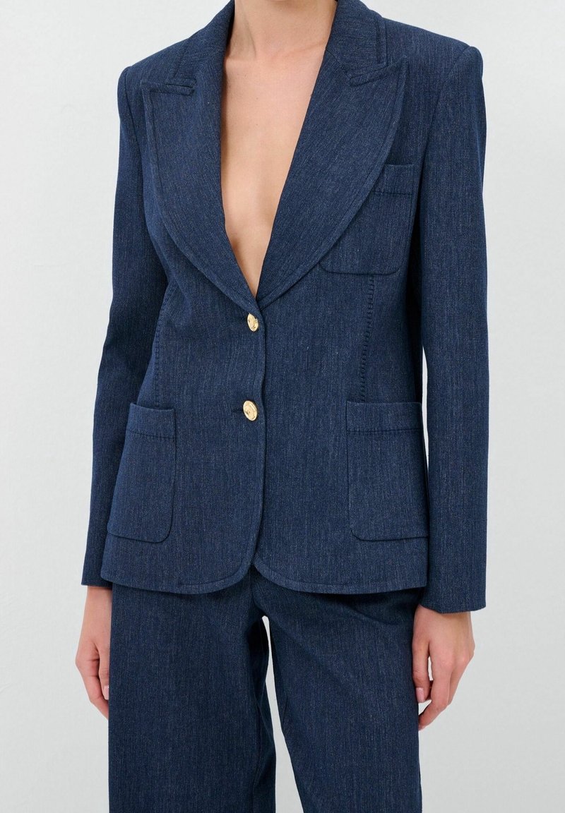 Blazer en denim bleu marine avec un col en V profond, deux poches frontales et des boutons dorés. Design structuré avec une coupe ajustée et une texture subtile.