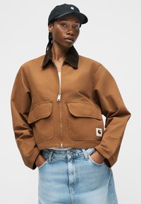 Persoană purtând o jachetă maro cu fermoar și buzunare cu clapetă, blugi din denim bleu deschis și o șapcă neagră, stând cu mâinile în buzunare.