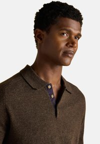 Man with short curly hair wearing a brown knit polo shirt with a navy buttoned placket, looking slightly to the right on a white background.