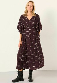 Langes, lila Kleid mit V-Ausschnitt, kurzen Ärmeln und geraffter Taille. Es hat ein leichtes, blumiges Muster in Creme und ist kombiniert mit schwarzen Schnürstiefeln.