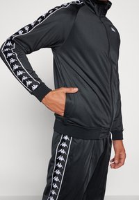 Personne portant un survêtement noir zippé avec des bandes blanches à logo courant le long des manches et du pantalon, une main dans la poche de la veste.