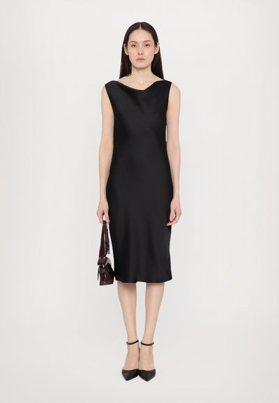 Vestido negro sin mangas en satén suave, con un escote ancho y curvado, y una silueta recta, combinado con unos tacones negros de punta y un bolso pequeño.
