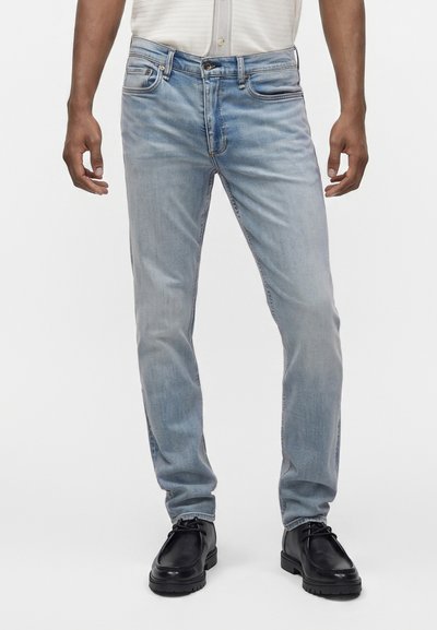 Άνδρας που φοράει ανοιχτό μπλε τζιν slim fit, σε συνδυασμό με μαύρα δερμάτινα παπούτσια με κορδόνια και μία λευκή πουκάμισο που είναι φορεμένη μέσα στο παντελόνι.