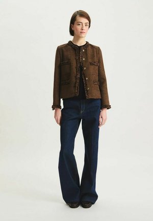 Veste marron texturée avec garniture décorative, quatre poches avant et boutons dorés, portée avec un pantalon en denim foncé évasé.