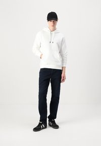 Hvid hættetrøje med lomme foran, kombineret med mørkeblå jeans og sorte sneakers med hvide striber. Modellen har en sort cap på.