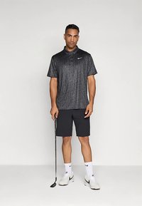 Černá vzorovaná polo košile s krátkými rukávy, logo Nike; v kombinaci s jednobarevnými černými šortkami. Stojí a drží golfovou hůl, má na sobě bílé ponožky a boty.