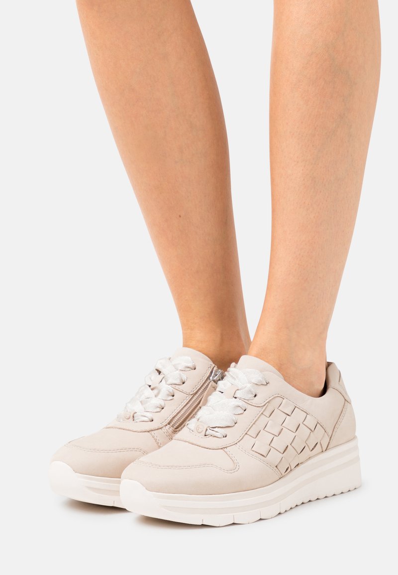 Baskets beige clair avec des accents tressés, présentant une surface texturée, un design à lacets et une fermeture éclair sur le côté. Semelle en caoutchouc avec légère élévation.