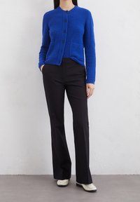 Modrý texturovaný svetřík s pěti černými knoflíky a dvěma předními kapsami, spárovaný s černými přizpůsobenými kalhotami a bílými zdobenými botami.