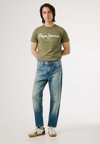 Camiseta de algodón verde oliva con el estampado blanco "Pepe Jeans London", combinada con jeans de mezclilla azul y zapatillas blancas con detalles negros.