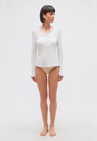 Γυναίκα με κοντά σκούρα μαλλιά, φοράει λευκό μακρυμάνικο πουκάμισο henley και μπεζ εσώρουχα, στέκεται ξυπόλυτη μπροστά σε λευκό φόντο.