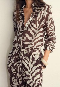 Chemise boutonnée à motif marron et crème, avec col et taille élastique, présentant une coupe décontractée et des manches longues.