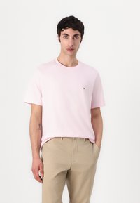 T-shirt rose clair à manches courtes avec un col rond, orné d'un petit logo sur la poitrine gauche, associé à un pantalon beige.