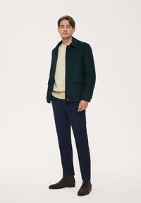 Grön jacka i ullblandning med framfickor, knäppning och krage, kombinerad med en cremefärgad tröja och marinblå byxor, avslutad med bruna stövlar.