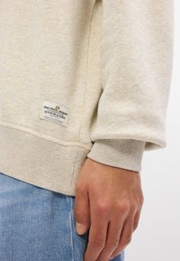 Ανοιχτό γκρι πλεκτό sweatshirt με ριγέ μανσέτες, που διαθέτει ετικέτα με λεπτομέρειες λογότυπου κοντά στο τελείωμα. Φοριέται με μπλε τζιν παντελόνι.