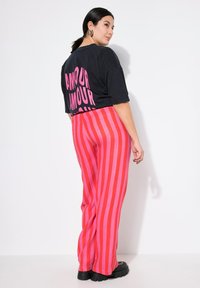 Zwarte oversized t-shirt met een felroze "AMOUR" opdruk op de achterkant, gecombineerd met breed gesneden broeken in rood-en-roze strepen en chunky zwarte schoenen.