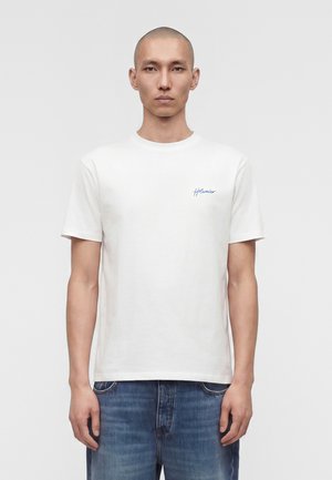 Άνδρας με κοντά μαλλιά, φοράει λευκή μπλούζα με μπλε γράμματα στο αριστερό στήθος και μπλε τζιν, στέκεται μπροστά σε ένα απλό γκρι φόντο.