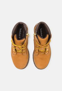 Hnědé semišové kotníkové boty se žlutými šňůrkami, polstrovaným límcem a bílým prošíváním. Zlacený doplněk na patě. Vyznačují se texturovaným povrchem.