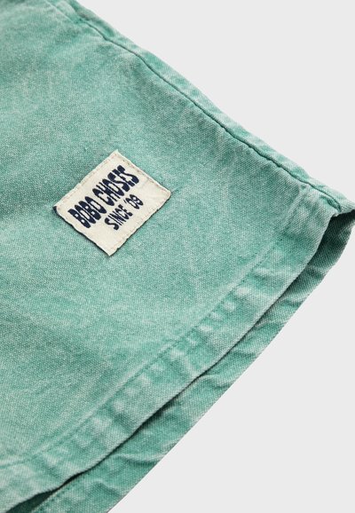 Tissu vert avec couture visible et une étiquette rectangulaire blanche indiquant "BOBBY CROSS SINCE '93" cousue près du bord.