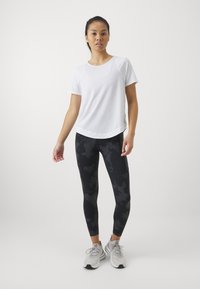 T-shirt atlético branco de mangas curtas, combinado com leggings pretas com um padrão de camuflagem discreto. Ténis cinzentos de corrida completam o visual.