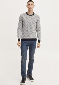 Grå strikkegenser med diamantmønster, svarte detaljer ved mansjettene og halsen, kombinert med blå jeans og svarte joggesko.