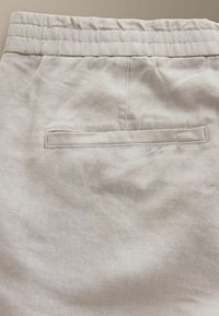 Pantalon en lin beige avec ceinture élastique et une poche arrière passepoilée, montrant la texture du tissu et les détails des coutures.