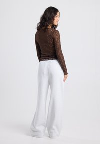 Vrouw met lang, donker haar, gekleed in een doorschijnende, bruine kanten top met lange mouwen en lichtgrijze, wijde broek, staand in een neutrale studio-omgeving.