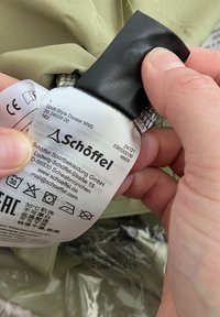 Etikett mit schwarzem Rand und weißem Hintergrund, das Pflegehinweise und das Markenlogo zeigt; von einer Hand über einem hellgrünen Stoff gehalten.