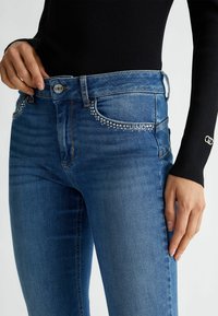 Primer plano de una persona que lleva jeans azules con detalles de strass en los bolsillos y una camiseta de manga larga negra con textura acanalada y un pequeño logotipo plateado en el puño.