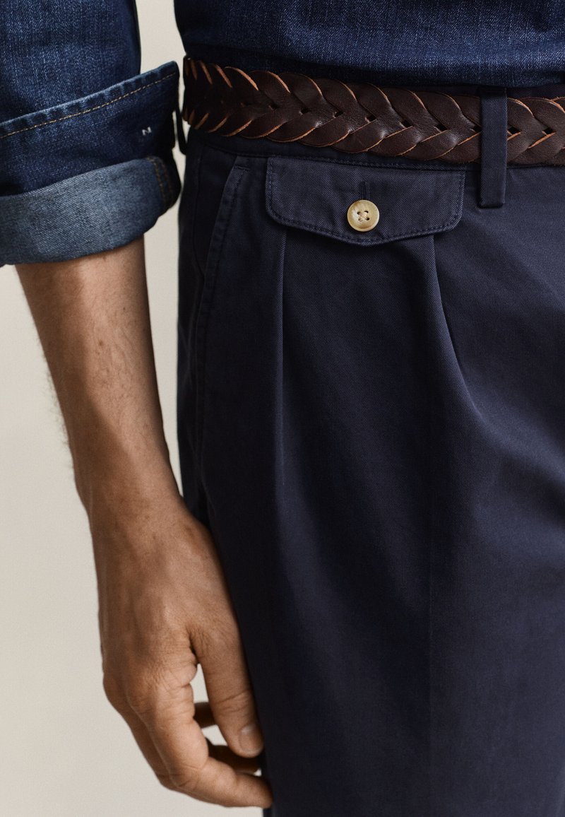 Primo piano di una persona che indossa pantaloni blu scuro con tasca a patta con bottone e cintura intrecciata in pelle marrone, mano appoggiata accanto alla gamba.