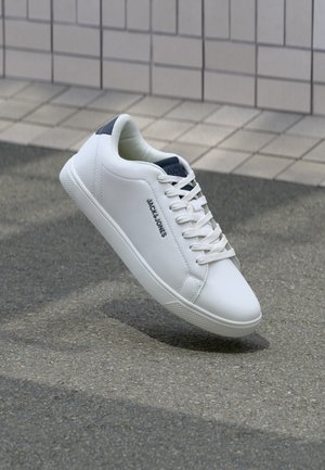 Zapatilla blanca de corte bajo con cordones, lengüeta trasera azul marino, marca "JACK & JONES" en el lateral, mostrada ligeramente inclinada sobre pavimento gris cerca de una pared de azulejos.