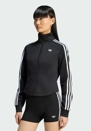 Donna che indossa una giacca da corsa Adidas nera con strisce bianche sulle maniche e pantaloncini neri abbinati con strisce bianche ai lati.