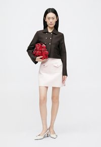 Bruine cropped jas met zilveren knopen, roze minirok met zakken aan de voorkant. Model houdt een kleine rode tas vast; witte sandalen met hak. Neutrale achtergrond.