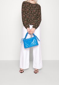 Sac à main bleu embossé crocodile avec une anse froncée, associé à un chemisier floral et un pantalon blanc à jambes larges, mettant en valeur un design contemporain.