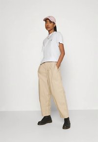 T-shirt blanc à manches courtes avec logo, pantalon beige ample, et bottines noires. Porte une casquette rose clair. Design simple et décontracté.