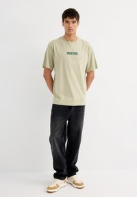 Camiseta de algodón verde claro con mangas cortas y texto impreso, combinada con jeans de mezclilla oscura y zapatillas beige con acentos en marrón.