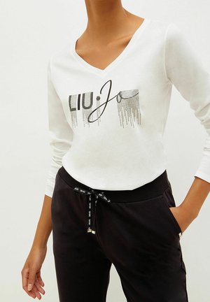 Personne portant un haut blanc à manches longues avec col en V et texte graphique noir, ainsi qu'un pantalon noir à taille à cordon avec la main dans la poche.