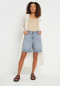 Cárdigan de ganchillo ligero sobre una camiseta sin mangas beige, combinado con pantalones cortos de denim azul y sandalias marrones con tachuelas decorativas.