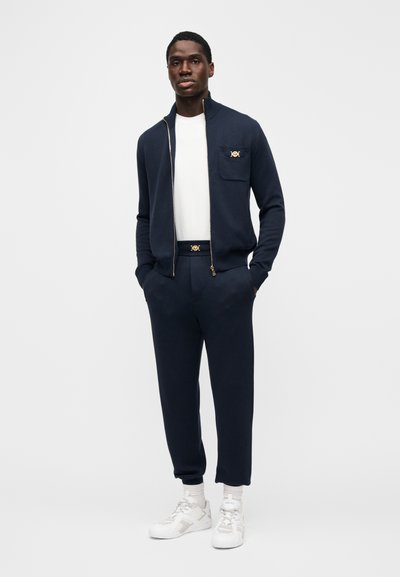 Marineblauw jack met ritssluiting en goudkleurig logo op de zak, gecombineerd met een bijpassende broek. Witte T-shirt en witte sneakers maken de outfit compleet.