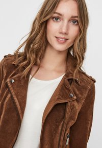 Bruine suède jacket met een ritssluiting aan de voorkant, schouderepauletten en zilverkleurige details, gecombineerd met een eenvoudig wit shirt eronder.